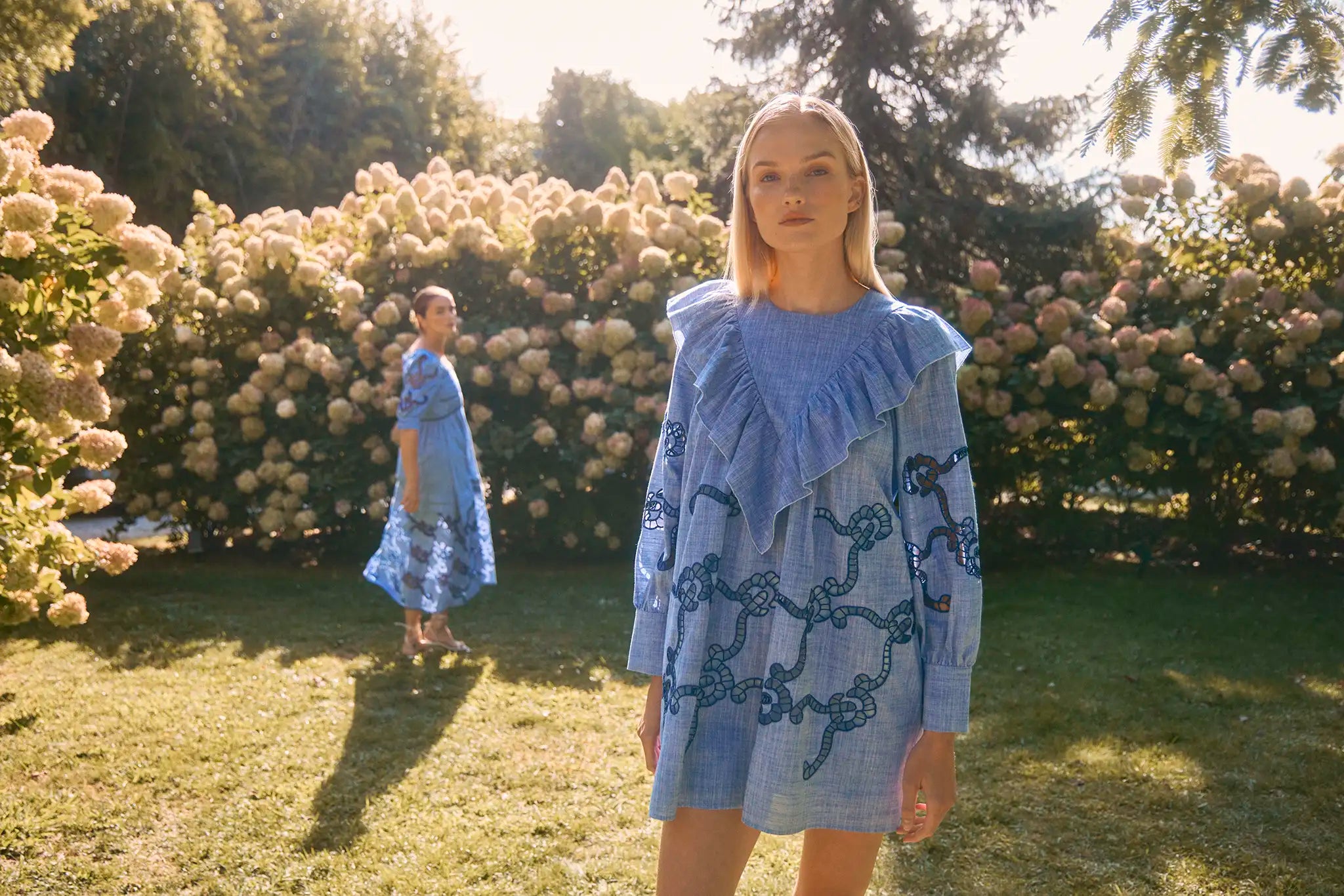Woman in a blue dress standing in a garden with floral bushes.