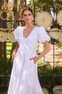 The Augusta Dress, white cotton dress with ruched V neckline, in front of a fence in the sun.