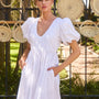 The Augusta Dress, white cotton dress with ruched V neckline, in front of a fence in the sun.