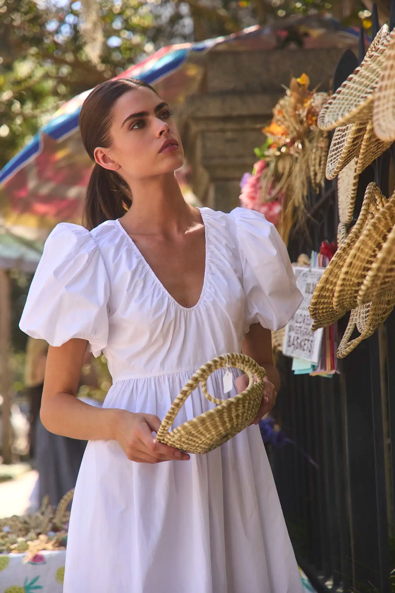 The Augusta Dress, white cotton dress with ruched V neckline, in front of a fence in the sun.